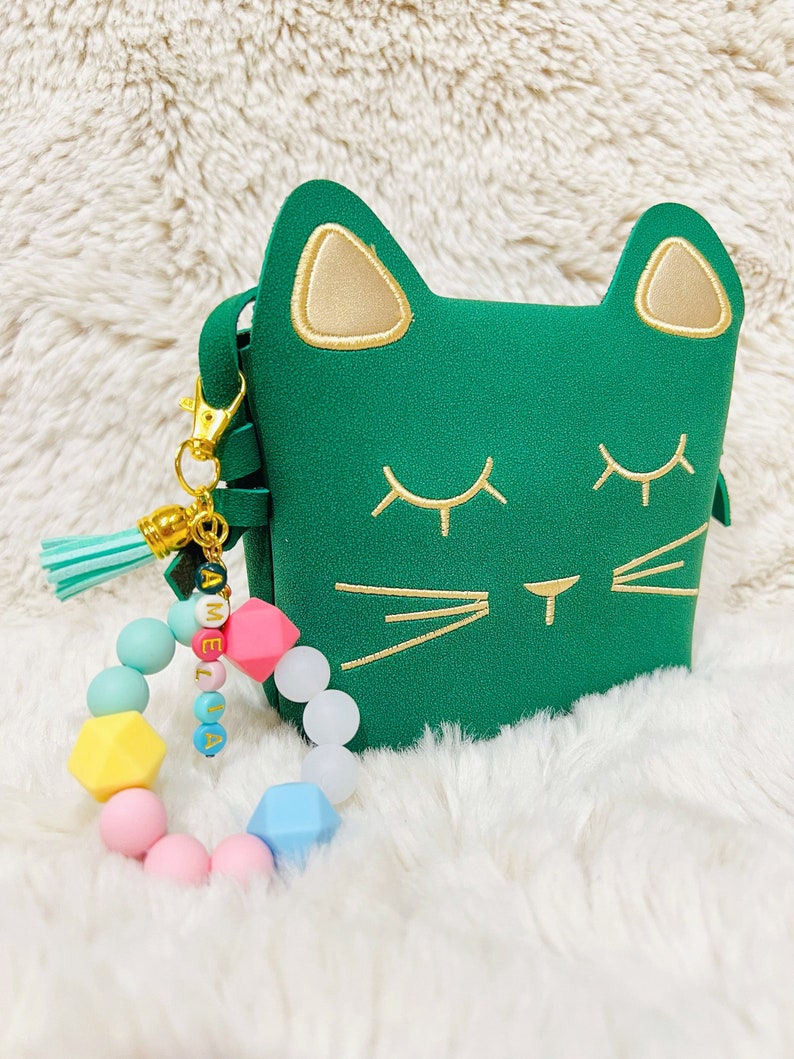 May include: A green cat-shaped purse with gold accents and a matching tassel keychain. The keychain also features a colourful beaded bracelet with the name "Amelia" spelled out.