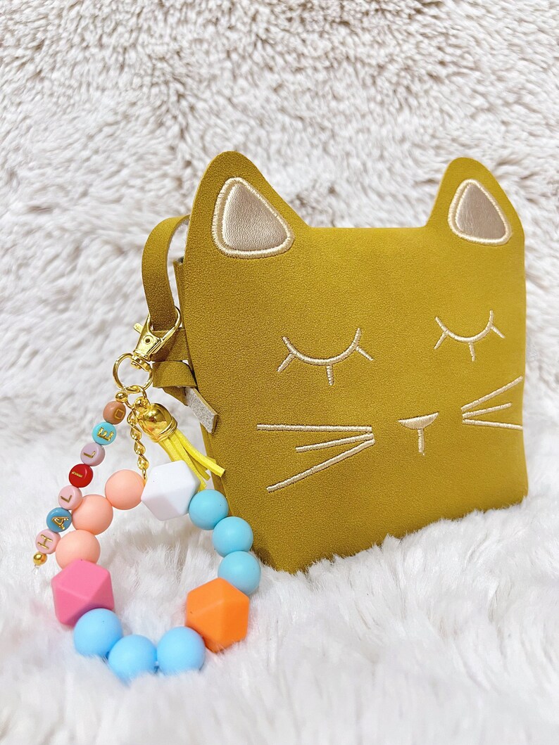 May include: A yellow cat-shaped purse with a gold chain and a colourful silicone beaded bracelet with the word "HALLO" spelled out.
