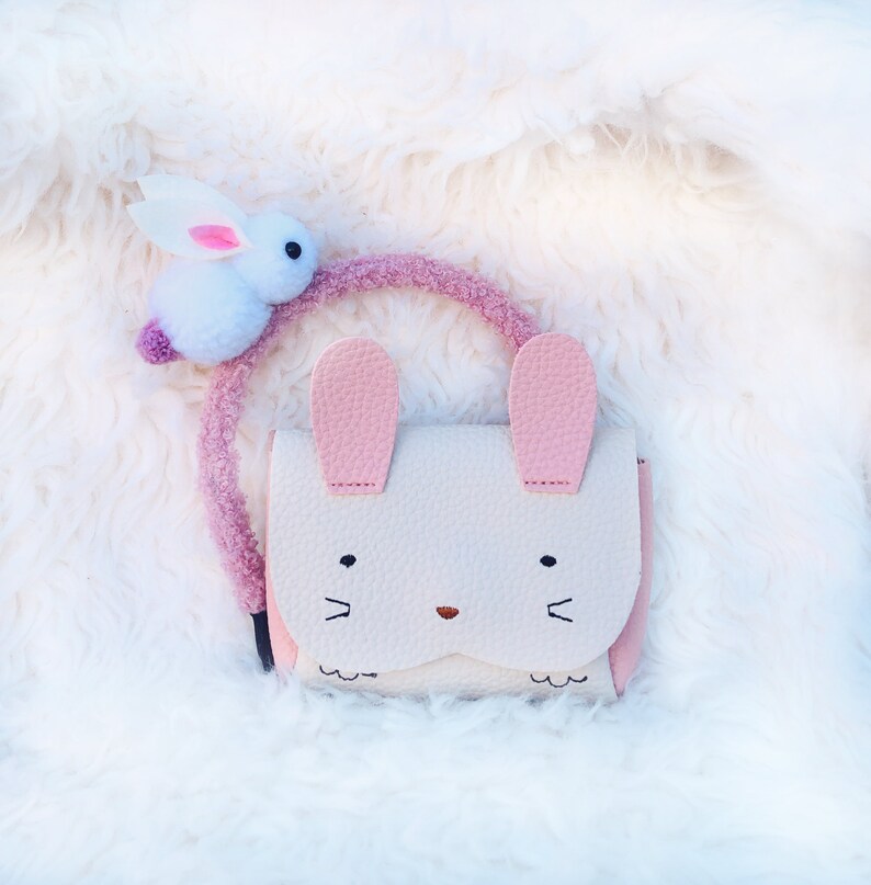 Pode incluir: Uma bolsa rosa e branca em forma de coelho com um amuleto de coelho branco felpudo preso &agrave; al&ccedil;a.
