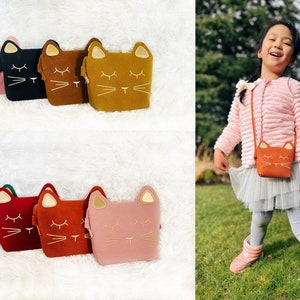 May include: Six different coloured cat-shaped purses with gold embroidery. The purses are lined up on a white fluffy surface. A young girl is wearing a pink sweater and a tulle skirt and is carrying a brown cat-shaped purse.