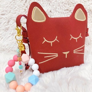 May include: A red cat-shaped purse with gold embroidered details and a gold tassel keychain. A colourful silicone bracelet with beads and the name "Olivia" spelled out in gold letters is also pictured.
