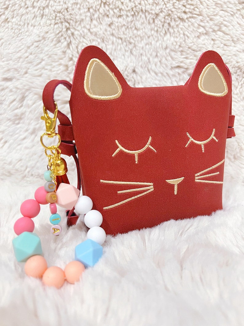 May include: A red cat-shaped purse with gold embroidered details and a gold tassel keychain. A colourful silicone bracelet with beads and the name "Olivia" spelled out in gold letters is also pictured.