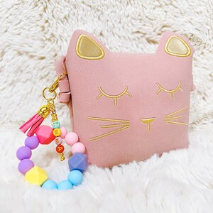May include: A pink cat-shaped pouch with gold embroidered details and a colourful beaded keychain with a pink tassel.