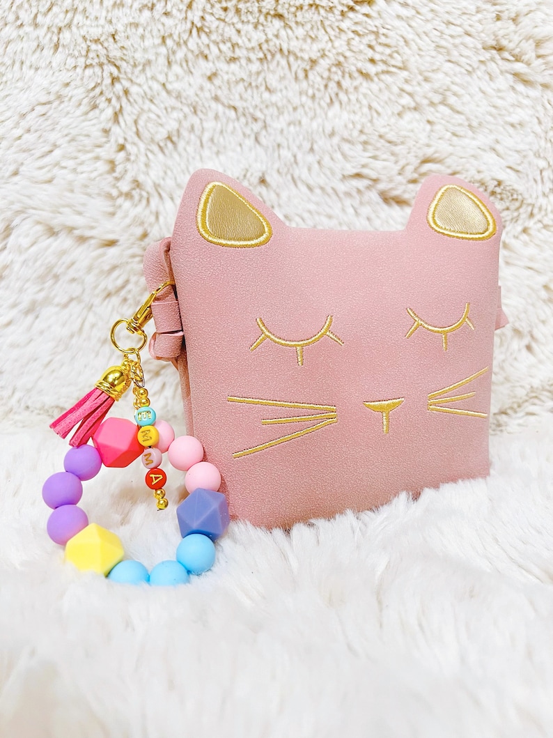 May include: A pink cat-shaped pouch with gold embroidered details and a colourful beaded keychain with a pink tassel.