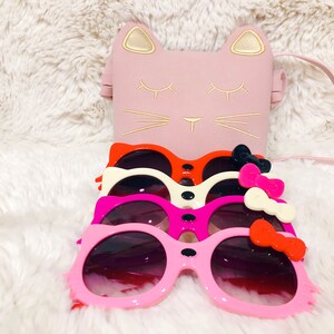 May include: Four pairs of pink, red, white and black cat-shaped sunglasses with bows. The sunglasses are stacked on top of each other. A pink cat-shaped purse with a gold trim is in the background.