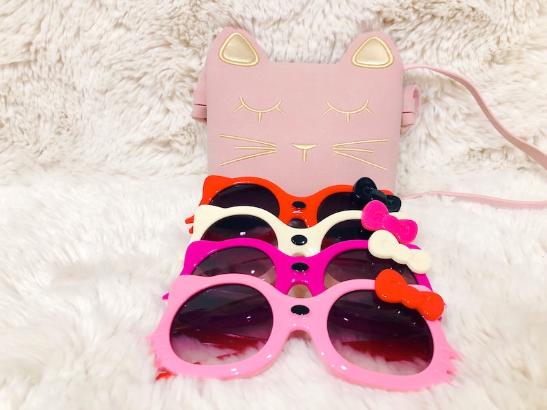 May include: Four pairs of pink, red, white and black cat-shaped sunglasses with bows. The sunglasses are stacked on top of each other. A pink cat-shaped purse with a gold trim is in the background.