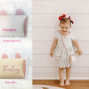 Pode incluir: Duas pequenas bolsas rosa e creme com orelhas de coelho. Uma bolsa tem o nome "Harper" bordado e a outra o nome "Hailey" em letras de ferro-on de glitter rosa.