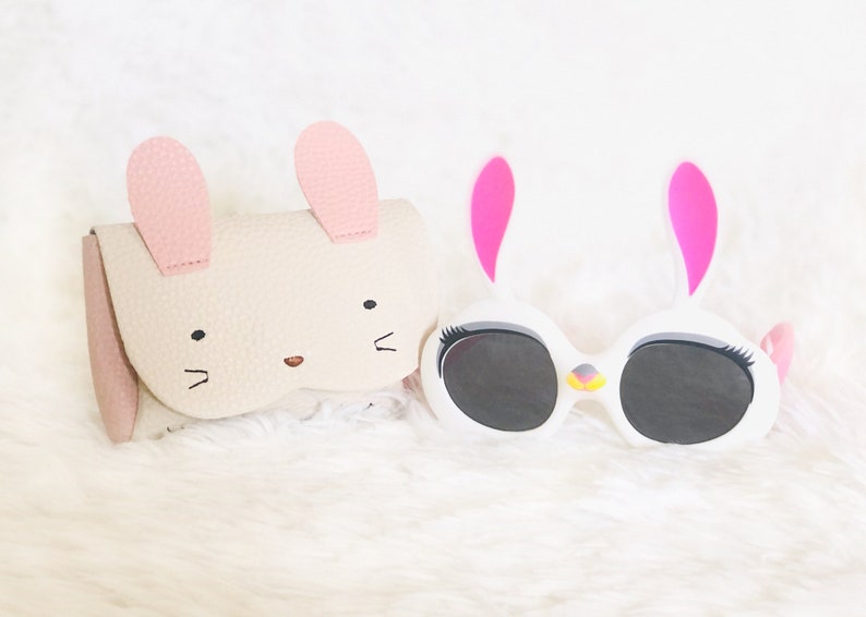 Pode incluir: Dois estojos de &oacute;culos de pel&uacute;cia em forma de coelho. Um &eacute; branco com orelhas rosa e c&iacute;lios pretos. O outro &eacute; bege com orelhas rosa e um nariz marrom.