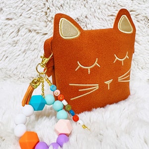 May include: A brown cat-shaped pouch with gold embroidered eyes and whiskers. A colourful silicone bracelet with a tassel is attached to the pouch.