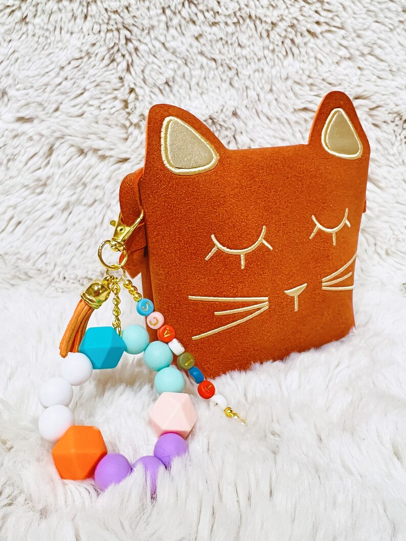 May include: A brown cat-shaped pouch with gold embroidered eyes and whiskers. A colourful silicone bracelet with a tassel is attached to the pouch.