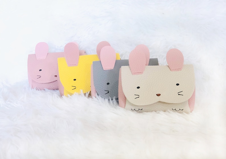 Pode incluir: Quatro pequenas bolsas coloridas em forma de coelhos com orelhas cor-de-rosa. As bolsas s&atilde;o feitas de couro sint&eacute;tico e t&ecirc;m olhos e narizes bordados. As bolsas est&atilde;o dispostas em fila sobre uma superf&iacute;cie branca e fofa.