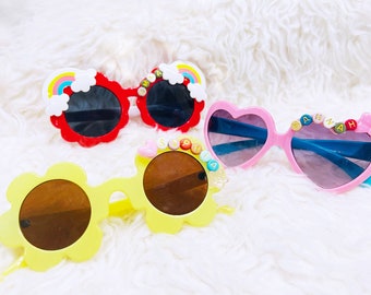 May include: Three pairs of colorful sunglasses with beaded embellishments spelling out names. The sunglasses are in the shapes of a flower, a heart, and a cloud.
