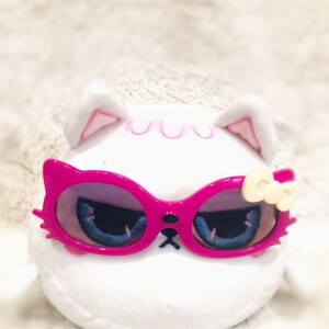 May include: White plush toy with pink ears and a pink bow. The toy is wearing pink sunglasses with a blue and white design.