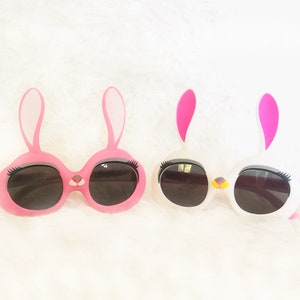 Pode incluir: Dois pares de &oacute;culos de sol em forma de coelho com arma&ccedil;&otilde;es rosa e brancas e lentes pretas. Os &oacute;culos t&ecirc;m c&iacute;lios e um pequeno detalhe de nariz.
