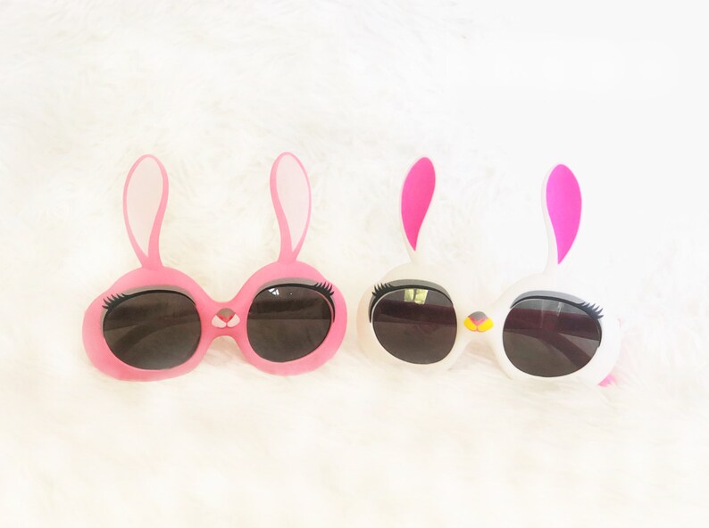 Pode incluir: Dois pares de &oacute;culos de sol em forma de coelho com arma&ccedil;&otilde;es rosa e brancas e lentes pretas. Os &oacute;culos t&ecirc;m c&iacute;lios e um pequeno detalhe de nariz.