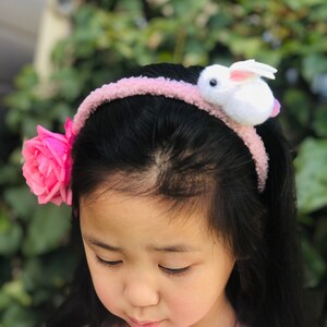 Pode incluir: Uma menina pequena usando uma faixa de cabe&ccedil;a rosa com um coelho branco e uma rosa rosa no lado.