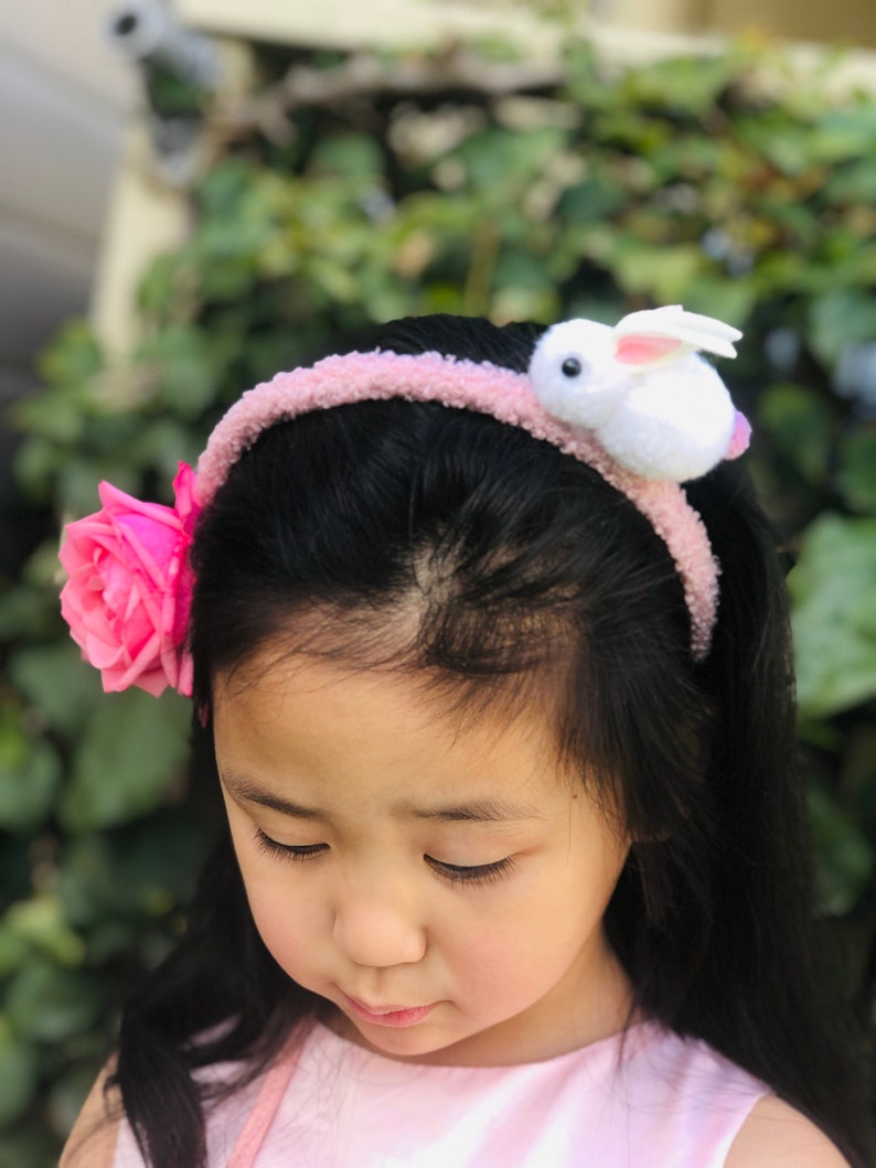 Pode incluir: Uma menina pequena usando uma faixa de cabe&ccedil;a rosa com um coelho branco e uma rosa rosa no lado.