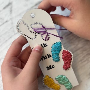 May include: A handmade white tag with the words "Walk With Me" and colorful embroidered footprints. The tag is being held by hands, with purple thread detailing the top and a hole for hanging.