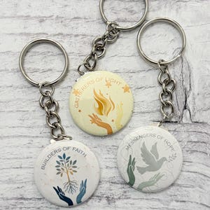 Puede incluir: Tres llaveros redondos con anillos y cadenas de metal plateado. Cada llavero presenta un diseño diferente con el texto: "Builders of Faith", "Gatherers of Light" y "Messengers of Hope". Los diseños incluyen un árbol, manos, una llama y una paloma.