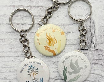 Pode incluir: Três chaveiros redondos com argolas e correntes de metal prateado. Cada chaveiro apresenta um design diferente com o texto: "Builders of Faith", "Gatherers of Light" e "Messengers of Hope". Os desenhos incluem uma árvore, mãos, uma chama e uma pomba.