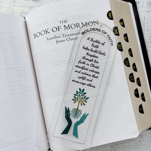Peut inclure: Un marque-page en acrylique transparent avec le texte "A Builder of Faith helps build God's kingdom through her faith in Christ, steadfast witness, and actions that uplift and encourage others." Le marque-page présente un motif d'arbre avec des mains et est placé dans un livre ouvert.
