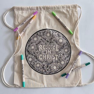 以下が含まれることがあります： ベージュのコットン製巾着型バックパック。中央に「REJOICE IN CHRIST」の文字と花や太陽のモチーフが描かれた円形の白黒ラインアートがプリントされており、周囲に5本のカラフルなアクリルペイントマーカーが配置されています。