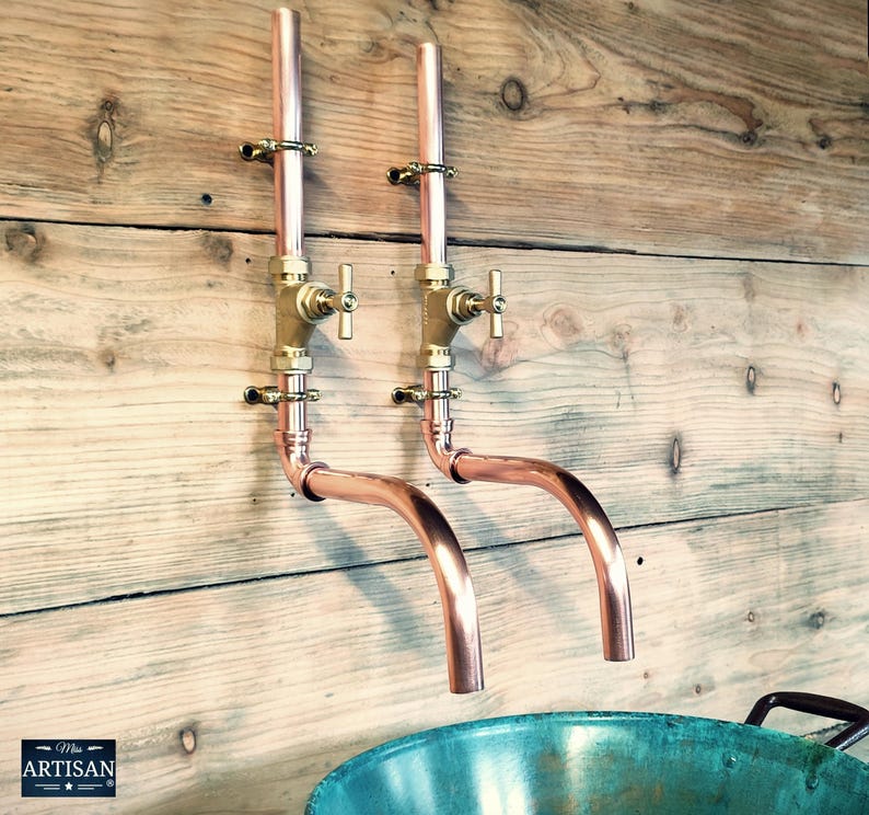 May include: Two copper wall-mounted faucets with brass handles and a curved spout. The faucets are mounted on a wooden wall and are connected to a copper pipe.