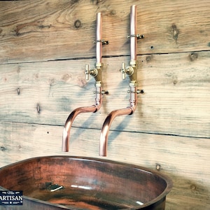 May include: Two copper wall-mounted faucets with brass handles and a copper oval sink. The faucets have a curved spout and are mounted on a wooden wall.