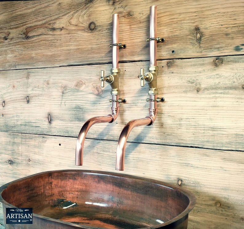 May include: Two copper wall-mounted faucets with brass handles and a copper oval sink. The faucets have a curved spout and are mounted on a wooden wall.