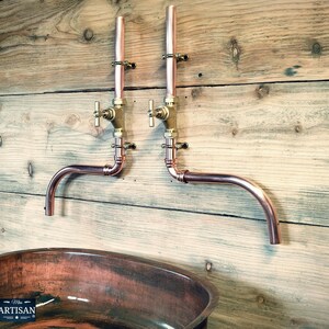 May include: Copper pipe wall mounted bathroom sink faucet with two brass valves. The faucet has a curved spout and is mounted on a wooden wall. The sink is a copper oval basin.