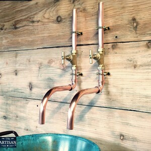 May include: Two copper wall-mounted faucets with brass handles and a curved spout. The faucets are mounted on a wooden wall.