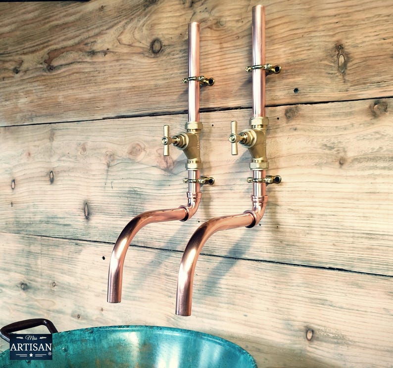 May include: Two copper wall-mounted faucets with brass handles and a curved spout. The faucets are mounted on a wooden wall.