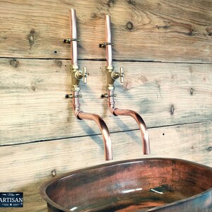 May include: Two copper wall-mounted faucets with brass handles and a copper oval sink. The faucets have a curved spout and are mounted on a wooden wall.