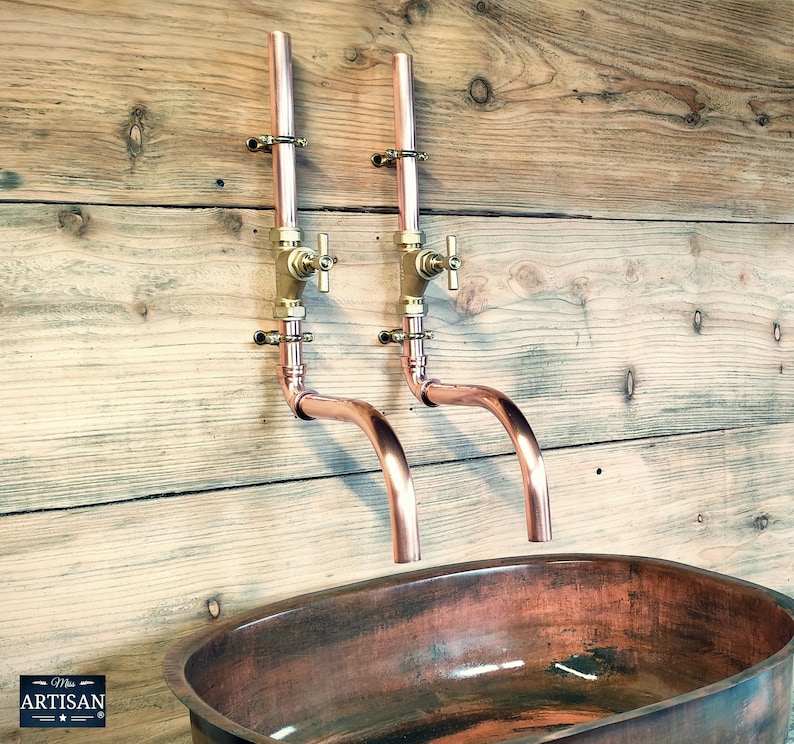May include: Two copper wall-mounted faucets with brass handles and a copper oval sink. The faucets have a curved spout and are mounted on a wooden wall.