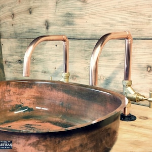Può includere: Lavabo da bagno in rame con due rubinetti in tubo di rame. Il lavabo è di forma ovale e ha una finitura rustica. I rubinetti sono fissati al muro con raccordi per tubi neri.