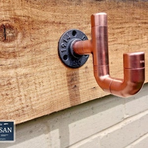 May include: Copper pipe coat hook with a black industrial pipe flange. The hook is mounted on a wooden surface.