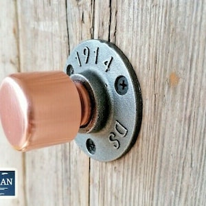 Könnte beinhalten: Ein kupferner Türknauf mit rundem, glattem Design. Der Knauf ist an einer schwarzen Metallflansch mit den Zahlen "1914" und den Buchstaben "DS" geprägt.