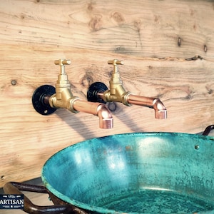 Può includere: Due rubinetti in ottone con tubi in rame sono montati su una parete di legno. I rubinetti hanno basi circolari nere e sono posizionati sopra un lavabo turchese. L'immagine ha un'estetica rustica e industriale.