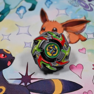 Op de afbeelding: Een kleurrijke tol met rode, groene en zilveren accenten, geplaatst tegen een achtergrond met stripfiguren. De tol heeft een centraal embleem met een drakenontwerp.