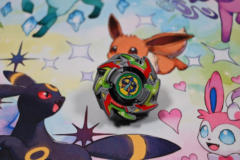 Op de afbeelding: Een kleurrijke tol met rode, groene en zilveren accenten, geplaatst tegen een achtergrond met stripfiguren. De tol heeft een centraal embleem met een drakenontwerp.