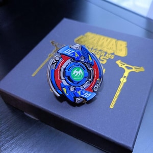 May include: A blue, red, and silver spinning top toy with the number "07" printed on it. The toy is on a gray box with gold accents and text. A gold tool is also visible.