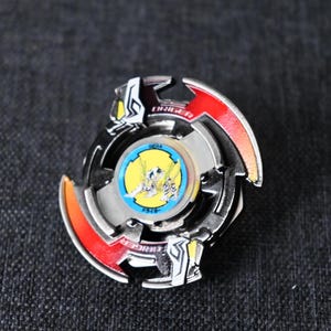 May include: A black, red and silver Beyblade with a yellow and blue centre. The Beyblade has the word "Engineer" printed on it.