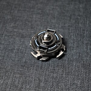 Op de afbeelding: Een zilveren metalen fidget spinner met een zwart midden en een zilveren ring rond het midden. De spinner heeft een getextureerd oppervlak en een reeks kleine, zilveren tanden rond de rand.
