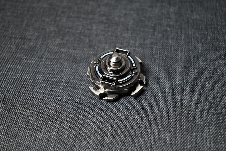 Op de afbeelding: Een zilveren metalen fidget spinner met een zwart midden en een zilveren ring rond het midden. De spinner heeft een getextureerd oppervlak en een reeks kleine, zilveren tanden rond de rand.