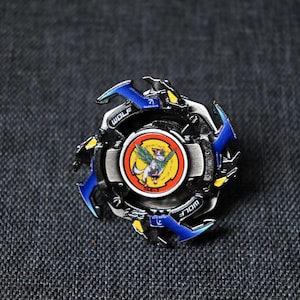Op de afbeelding: Een blauwe en zwarte metalen Beyblade met een wolfontwerp en een gele en rode ronde afbeelding van een wolf met vleugels in het midden. De Beyblade ligt op een grijze stoffen achtergrond.