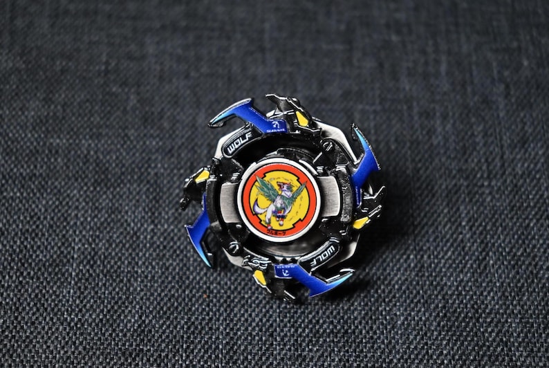 Op de afbeelding: Een blauwe en zwarte metalen Beyblade met een wolfontwerp en een gele en rode ronde afbeelding van een wolf met vleugels in het midden. De Beyblade ligt op een grijze stoffen achtergrond.