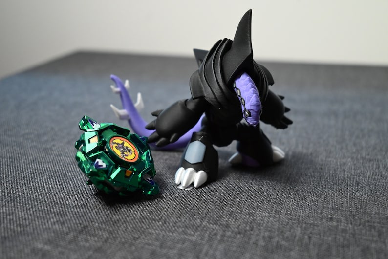 Puede incluir: Una peonza verde y negra con un s&iacute;mbolo amarillo y negro en el centro. La peonza est&aacute; al lado de un monstruo de juguete negro y morado con una aleta puntiaguda en la cabeza.