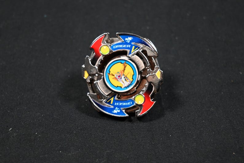 May include: A silver, blue, red, and yellow spinning top toy with the word "ORIGER" printed on it. The toy has a circular design with a central image and is set against a dark background.