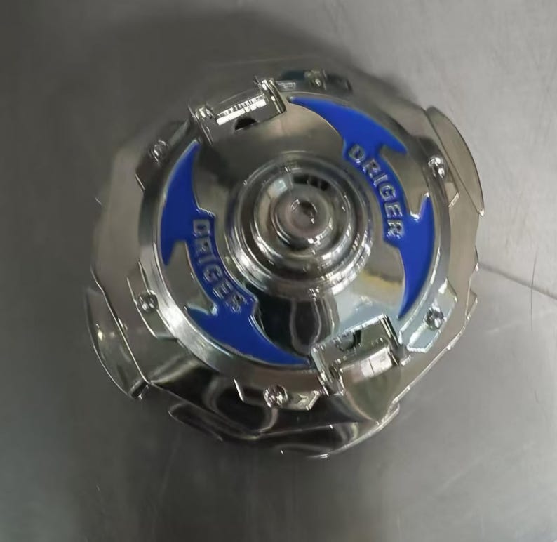 May include: A shiny, silver toy with blue accents and the word "DRIGER" printed on it. The toy has a circular shape with a complex design, featuring multiple layers and textures. The overall appearance is metallic and modern.