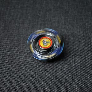 Op de afbeelding: Een draaiend speelgoed met een zwart, blauw en geel ontwerp. Het speelgoed heeft een transparante kunststof buitenlaag en een metalen binnenlaag.